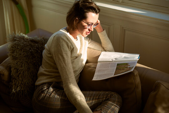 Woman reading a magazine on a sunlit couch, wearing glasses and a cozy sweater. Belgium