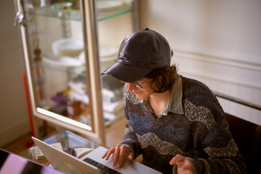 Person in a cozy sweater and cap using a laptop at a home desk setting Belgium