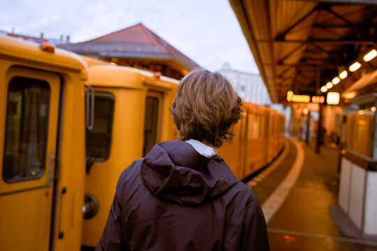 A person with brown hair waits at a train platform with yellow trains and an urban setting. Germany