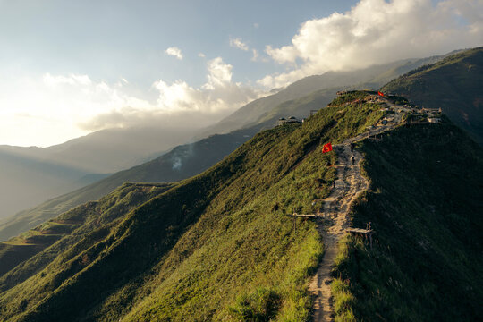 A winding path leads up a lush green mountain under a partly cloudy sky at sunset. Mountains of Ta Xua, Bac Yen District, Son La, Vietnam