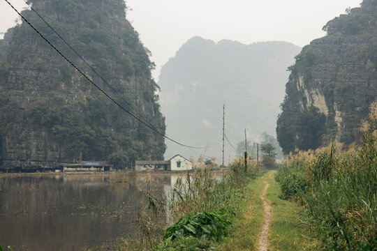 A narrow path leads through lush greenery beside a calm lake, flanked by large limestone cliffs. Hoa Lu District, Ninh B&Iuml;nh, Vietnam