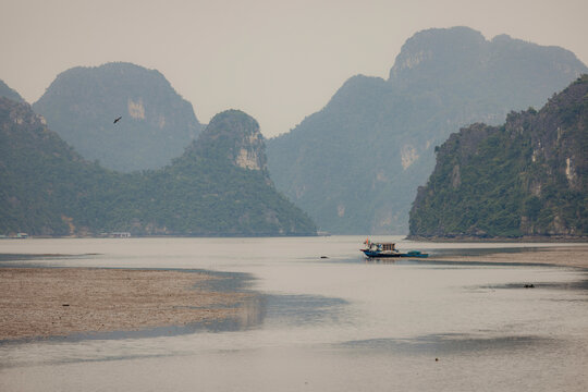 A lone boat on a vast, serene lake surrounded by towering green hills under an overcast sky. Cat Ba, Vietnam
