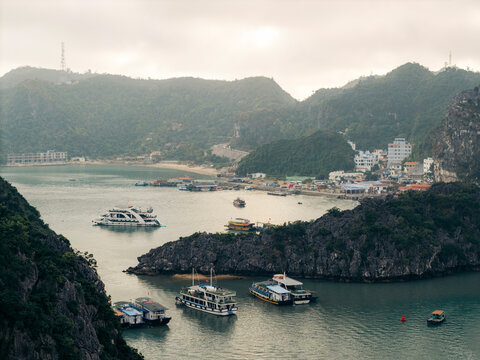 Boats sail through a bay surrounded by hills with a harbor town in the background. Lan Ha Bay, Cat Ba, Vietnam