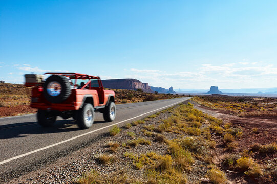 Red off-road vehicle drives along a deserted highway through a rocky desert landscape. Monument Valley, USA