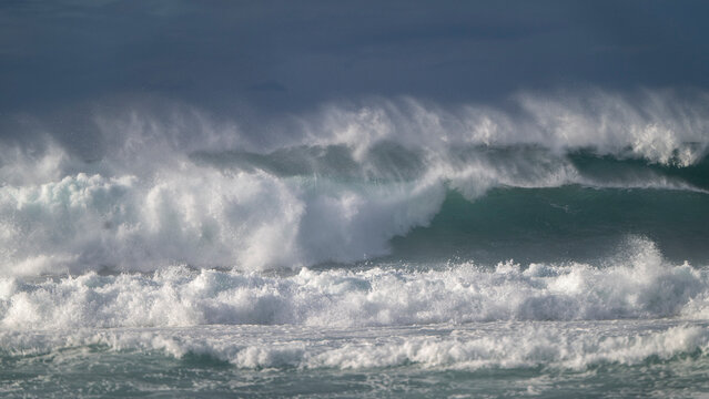 Powerful ocean waves crash under a dark, cloudy sky with white spray in the air. Paia, Hawaii, USA