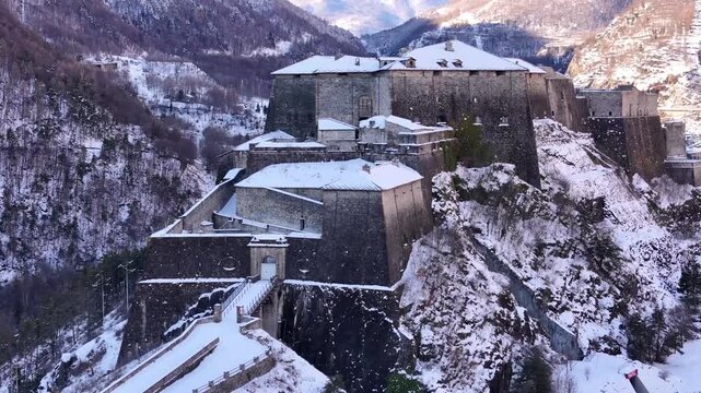 Aerial view of Forte di Exilles, a stone fortress covered in snow perched atop a mountain, contrasting with the bare trees, Exilles, Piemonte, Italy.