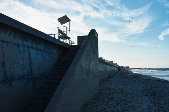Concrete seawall and stairs leading to a lifeguard chair under a blue sky with seagulls. Marshfield, MA, USA