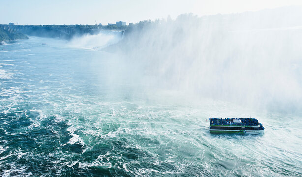A boat full of tourists approaches a massive waterfall under a misty sky. Niagara Falls, Ontario, Canada