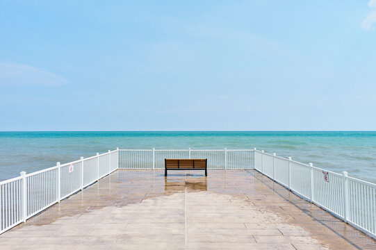 A lone wooden bench sits at the end of a pier overlooking a calm, blue ocean under a clear sky. Lake Michigan, Illinois, USA