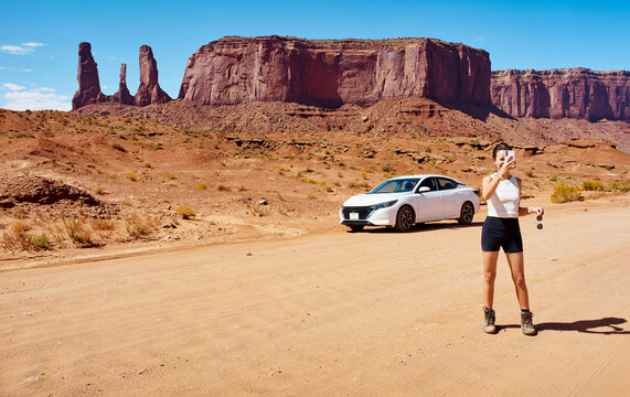 Young woman with skateboard on desert road with red rock formations and white car under clear sky. Monument Valley, Arizona, USA