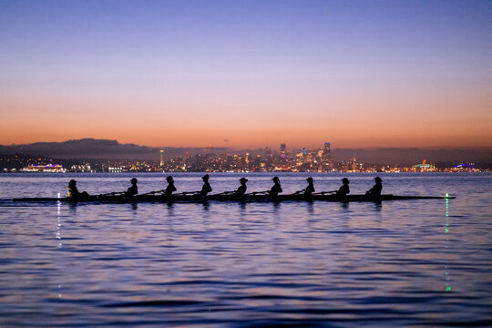 Rowing team in a boat silhouette against a city skyline at sunset on calm water. WA, USA