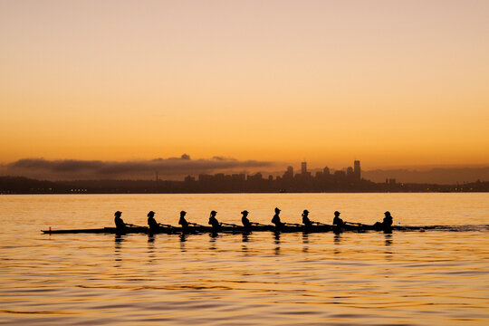 A rowing team glides on calm water at sunrise with a city skyline in the background. WA, USA