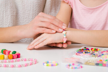 Mother hands putting bracelet on little child girl wrist on white table at home room. Creating beaded jewellery from different colorful plastic parts. Closeup. Front view.