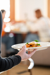A server holds a white plate with a gourmet seafood dish featuring scallops and shrimp in a restaurant setting.