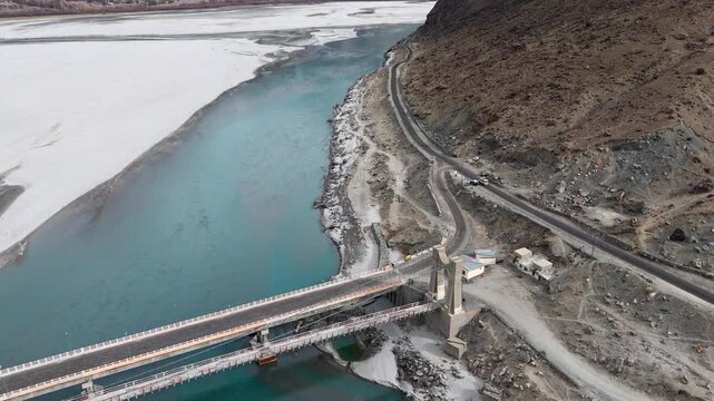 Aerial view of the bridge connecting the road to a partially frozen river with a road running along the side of a mountain, Daghoni Balgar, Gilgit Baltistan, Pakistan.
