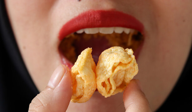 Close up of an Asian Muslim woman wearing hijab and red lipstick eating a crunchy corn snack, focusing on texture, snack craving, bold makeup, and modern lifestyle concept.