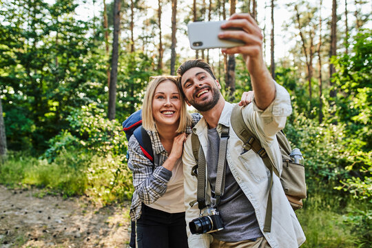 Young couple taking selfie while hiking in forest together during sunny summer day