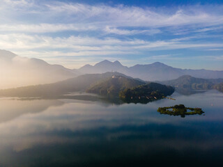 Aerial early morning view of Sun Moon Lake in Taiwan, with calm blue water surrounded by misty mountains and forested hills, capturing a serene atmosphere just after sunrise.

