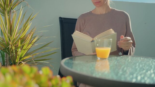 Solitary moment at home. Woman read and drink fresh orange juice alone in quiet terrace. Concept of solitude, introvert lifestyle, calm and peaceful indoor atmosphere.Copy space for text.