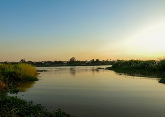 Obraz premium Wide tranquil river reflecting the soft golden light of sunset, framed by lush green reeds and rustic village dwellings under a peaceful dusk sky.