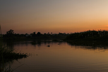 Quiet water surface reflecting dramatic golden hour sunset light over dark tropical forest and rural homes, creating a peaceful, tranquil atmosphere.