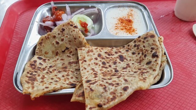 Pan shot of aloo paratha or flatbread served with creamy curd, onions, and crisp cucumber slices