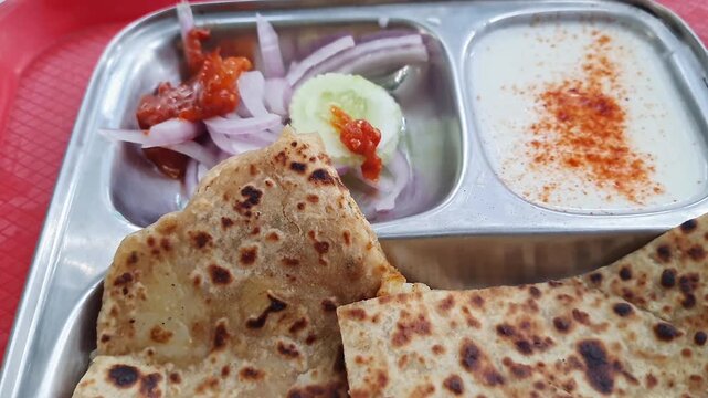 Pan shot of aloo paratha or flatbread served with creamy curd, onions, and crisp cucumber slices