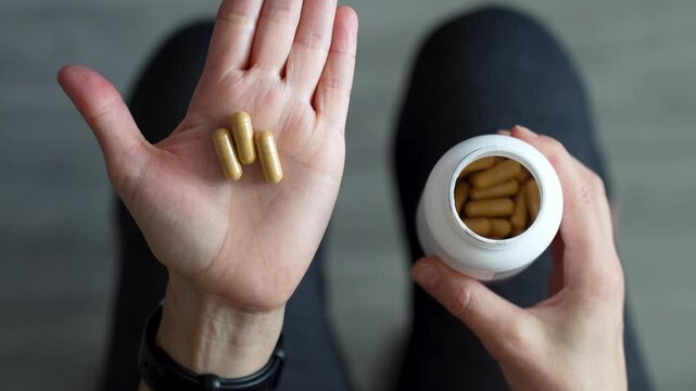 Top View of Woman Pouring Supplement Capsules Into Palm. A woman pours yellow medicine capsules from a white plastic bottle into her hand. Healthy habits, pharmaceutical treatment, and vitamin intake