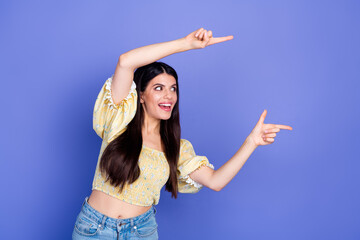 Young woman points with both hands in bright yellow blouse and jeans against a purple background