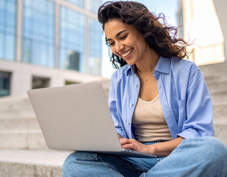 Junge Frau sitzt lachend mit ihren Laptop auf einer Stufe in der Stadt