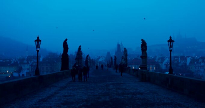Quiet winter morning on Charles Bridge in Prague with only a few people crossing. Cold blue atmosphere and soft dawn light create a peaceful and cinematic city scene.