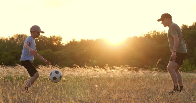 Father and son play soccer outdoors. Parent teaches kid soccer skills on field at sunset. Happy family enjoys active game. Boy kicks ball during soccer training in meadow evening light.