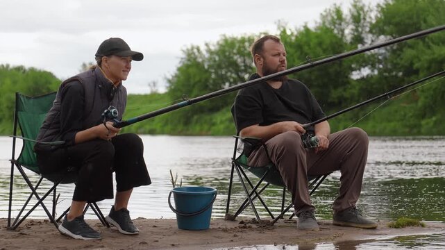 Man and woman enjoying a quiet day fishing together, sitting on folding chairs by the river with their rods in the water, relaxing during a weekend getaway in the beautiful countryside