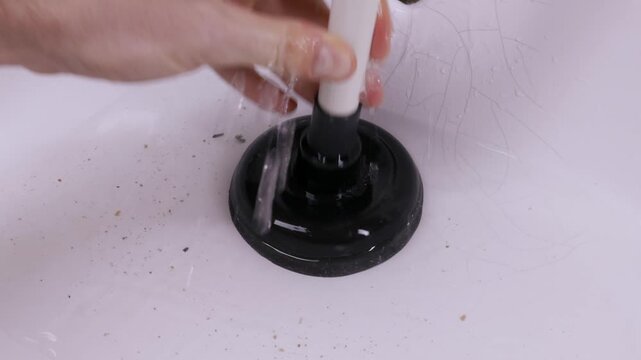 A male hand pumps a black plunger over a stainless steel drain to unclog a white sink filled with water and debris. This represents routine household maintenance and plumbing repair.