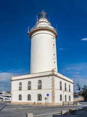 A tall white lighthouse rises from a stone base beside a quiet coastal street under a clear blue sky, Malaga, Spain