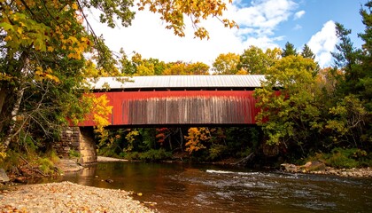 A vibrant autumn landscape featuring a wooden covered bridge spanning a flowing river. Surrounded by colorful fall foliage under a partly cloudy sky
