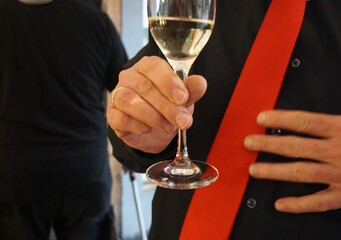 Stehender Mann mit schwarzem Hemd und roter Krawatte h&auml;lt Sektglas in Hand auf Feier in Raum