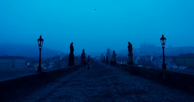 Quiet winter morning on Charles Bridge in Prague with only a few people crossing. Cold blue atmosphere and soft dawn light create a peaceful and cinematic city scene.