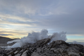 Sunset over the Atlantic Ocean, rocky shore covered by crashing waves, creating a dense wall of spray haze in dirty gray and golden tones, a fantastic shot of nature © Aliaksandr