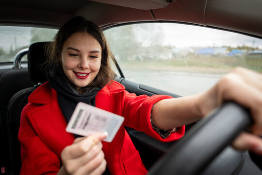 A young, attractive girl sits in a car, joyfully holding her new driver's license, celebrating passing her driving test.