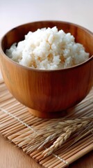 White rice in a wooden bowl rests on a bamboo mat with ears of wheat beside it against a gray background in a close-up shot