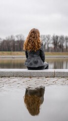 Fototapeta premium Woman with curly red hair sitting on the edge of a stone pier, gazing at the still water, with reflections visible and trees in the background