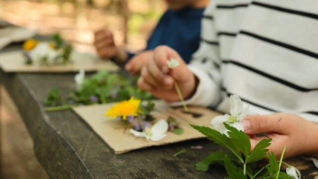 Children's hands crafting nature art with wildflowers and green leaves on a wooden table