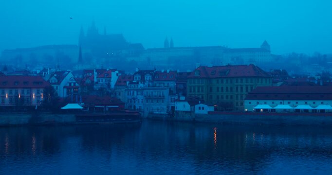 Wide winter view of Prague Old Town with Prague Castle in the background during blue hour. Moody fog and cold early morning light create a cinematic European cityscape.