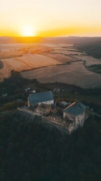 Tocnik Castle stands majestically atop a hill, showcasing its medieval ruins. The golden hues of dusk illuminate the castle's towers and surrounding landscape during a serene summer evening.