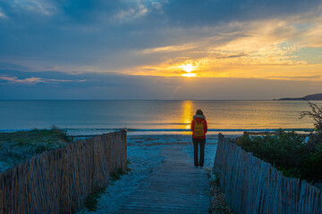 Woman Standing on Beach Path at Sunset with Calm Sea and Clouds