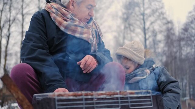 Gray haired grandfather and child by grill roasting sausages on charcoal brazier, bundled in winter coats and scarves on snowy ground. Hands warmed over glowing embers, smoke. Family outdoor barbecue.