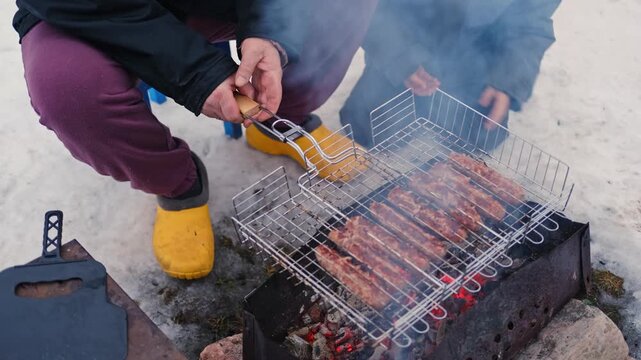 Gray haired grandfather and child by grill roasting sausages on charcoal brazier, bundled in winter coats and scarves on snowy ground. Hands warmed over glowing embers, smoke. Family outdoor barbecue.