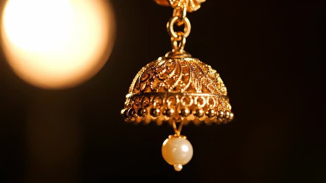 Close Up Of An Intricately Designed Golden Jhumka Earring With A Pearl Drop Isolated On A Dark Blurred Background With Soft Golden Light Bokeh