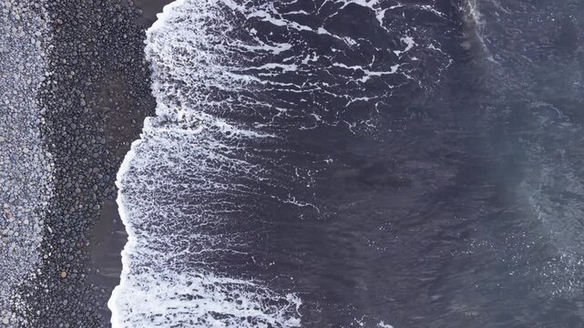Top-down aerial drone footage presenting a close-up overhead view of ocean waves and foam patterns interacting with a pebble shoreline.